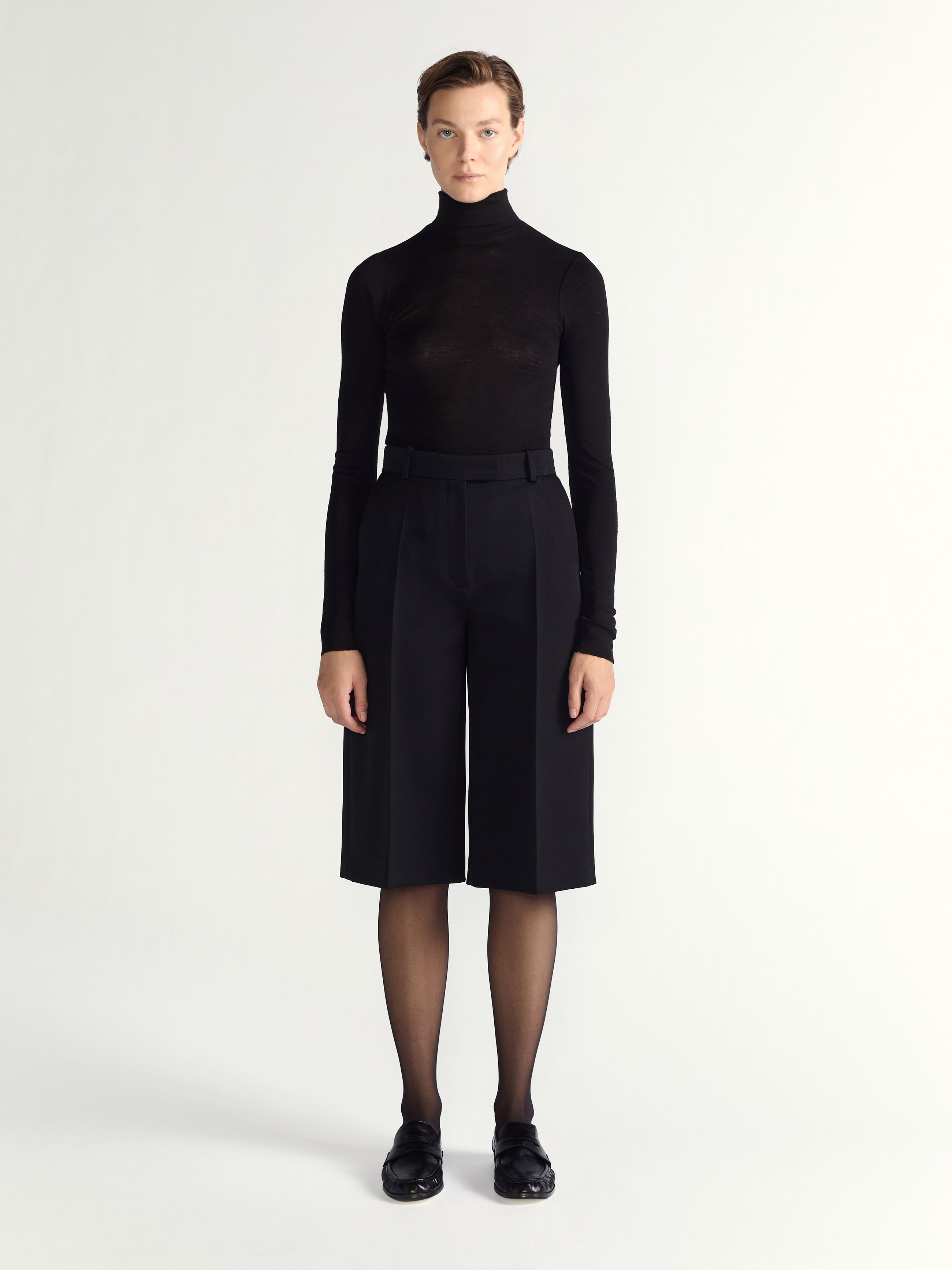 Person wearing a black turtleneck and black high-waisted shorts on a white background