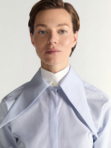 Person wearing a light blue striped shirt with a white collar against a plain background