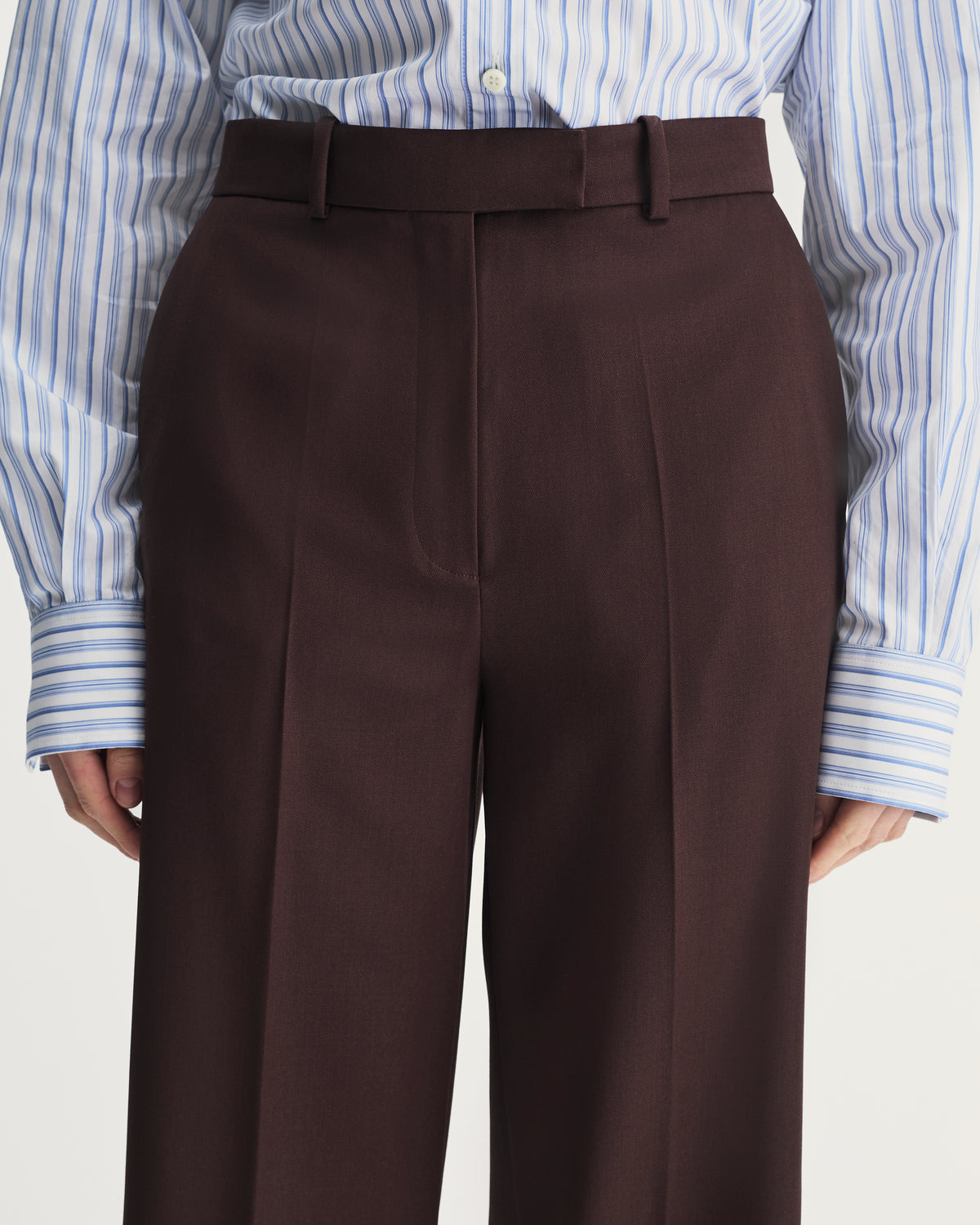 Brown pleated pants worn with a blue and white striped shirt on a light gray background