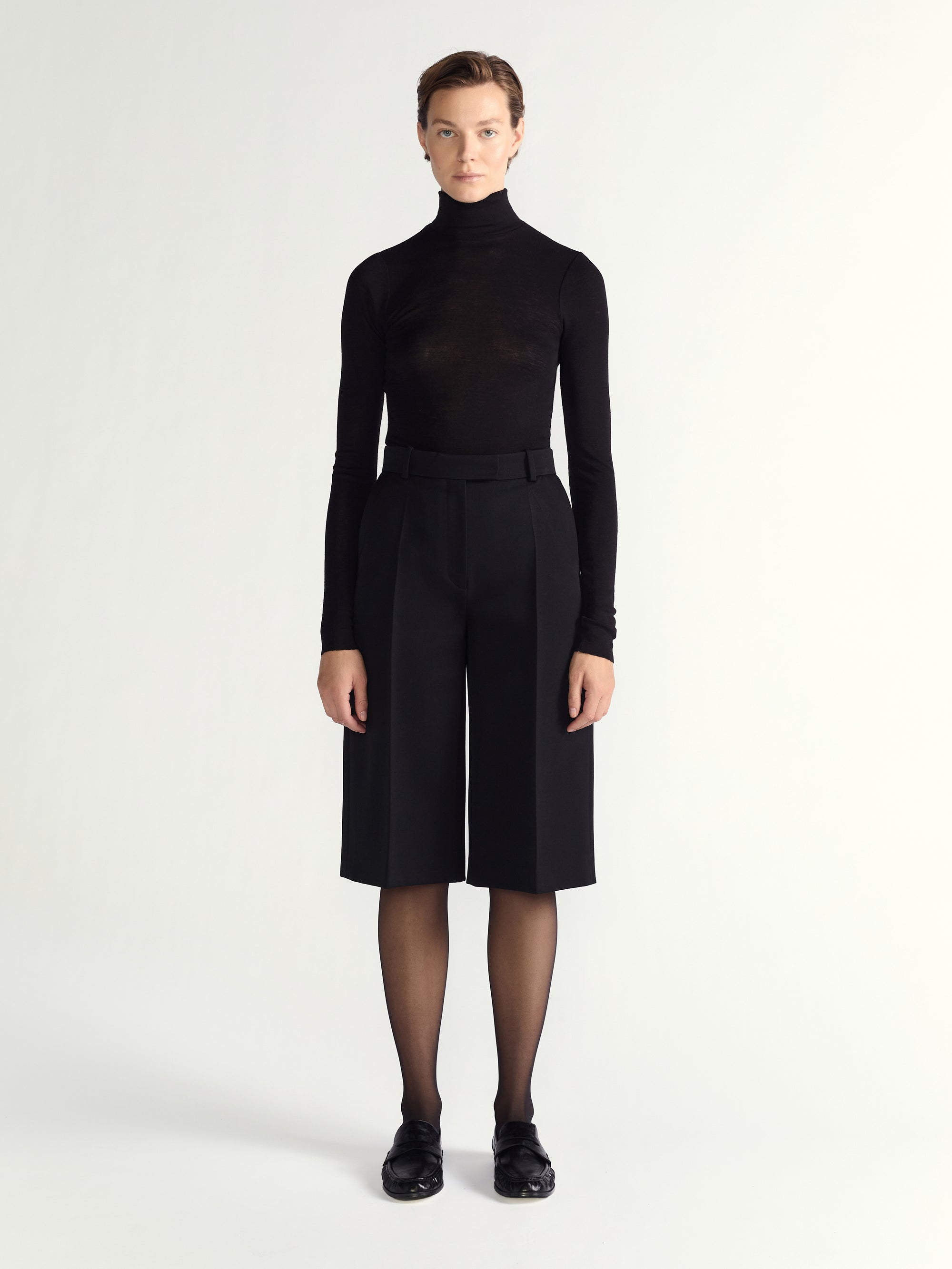 Person wearing a black turtleneck and black high-waisted shorts on a white background