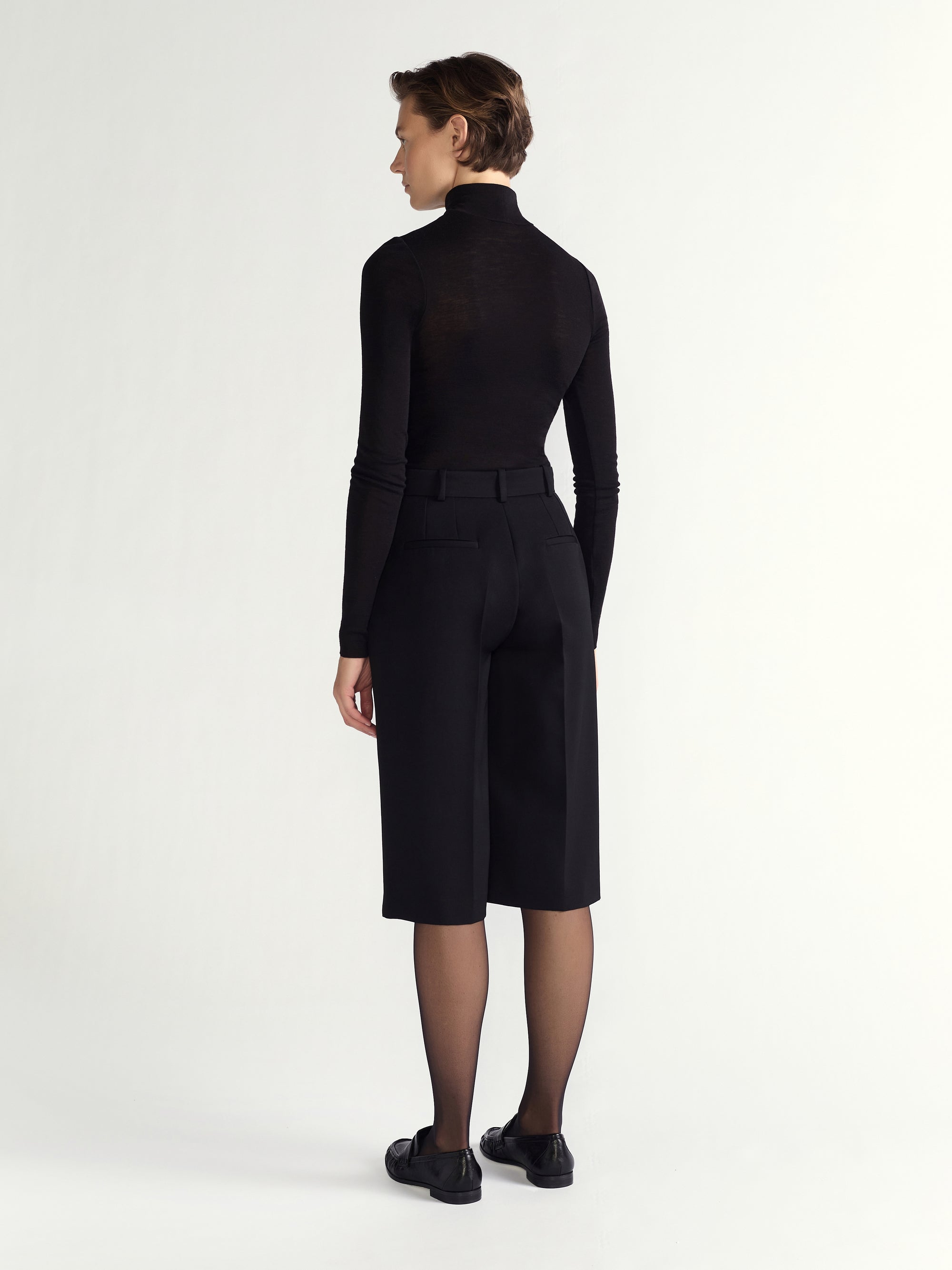 Person wearing a black outfit with a high collar and black shoes on a white background