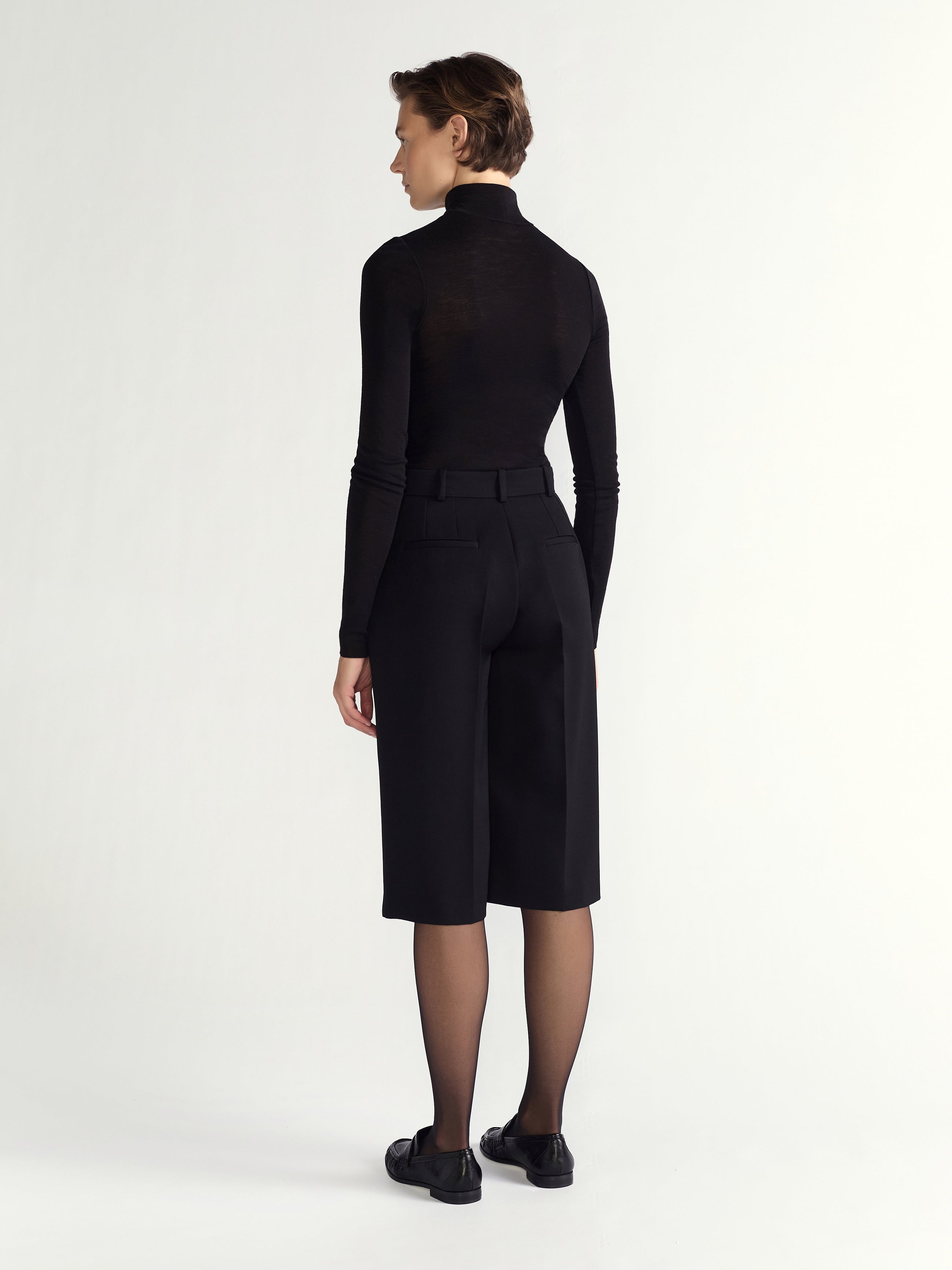 Person wearing a black outfit with a high collar and black shoes on a white background